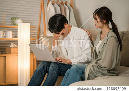 A wife looks anxiously at her husband working on a computer on the sofa in the living room (overtime, taking work home, hidden overtime) A wife looks anxiously at her husband working on a computer on the sofa in the living room (overtime, taking work home, hidden overtime) 138097409