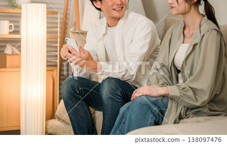 Young Asian couple chatting while looking at smartphone on the sofa in the living room at home Young Asian couple chatting while looking at smartphone on the sofa in the living room at home 138097476