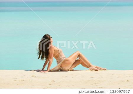 A beautiful tanned girl with an athletic physique in a swimsuit is siting on the beach on a sunny day in the Maldives, she is happy and posing 138097967