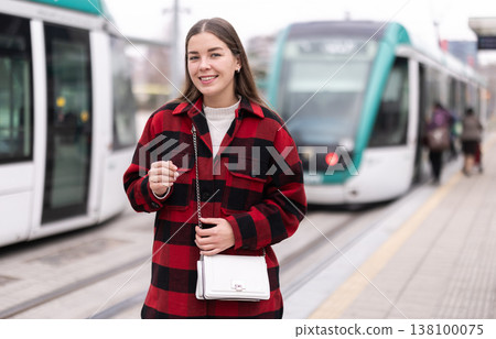 Girl is standing on street platform, waiting for tram to arrive Girl is standing on street platform, waiting for tram to arrive 138100075