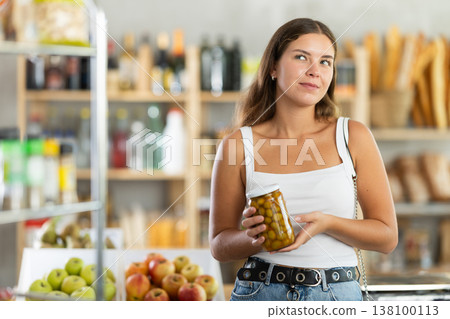 Young female shopper carefully choosing jar of canned olives in grocery supermarket 138100113