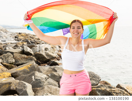 Portrait of a playful girl holding an LGBT flag in her hands on the beach on sunny day 138100190