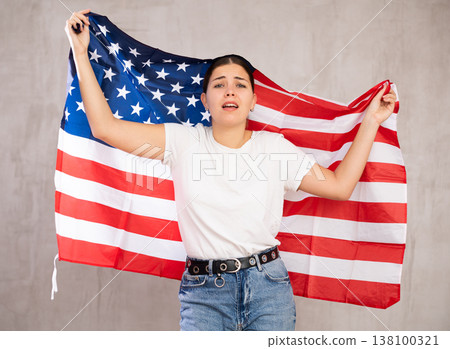 Sad young woman holding USA flag against unicoloured background Sad young woman holding USA flag against unicoloured background 138100321