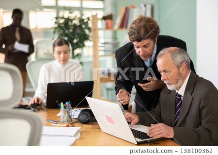 Man leaned over to seated mature male employee, helps with work and looks at laptop screen 138100362