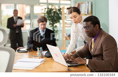Woman leaned over to seated African man employee, helps with work and looks at laptop screen 138100384