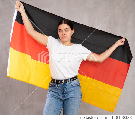 Serious young woman holding national flag of Germany on gray wall background Serious young woman holding national flag of Germany on gray wall background 138100388