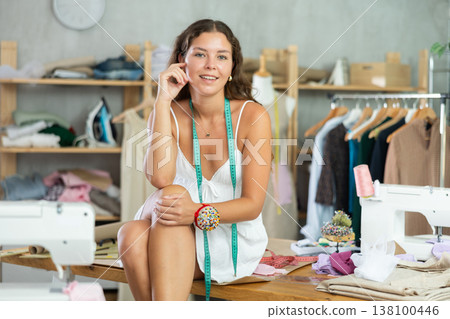 Playful young seamstress posing on table during break in atelier Playful young seamstress posing on table during break in atelier 138100446