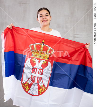 Cheerful patriotic young woman holding flag of Serbia against gray background indoors 138100548