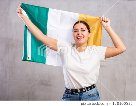 Excited young woman holding state flag of Ireland against gray wall background indoors Excited young woman holding state flag of Ireland against gray wall background indoors 138100553