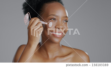 Facial roller, woman and smile in face massage in skincare or beauty, health and wellness or dermatology. Happy black person, skin and cosmetics with glow, care and tool in studio by gray background Facial roller, woman and smile in face massage in skincare or beauty, health and wellness or dermatology. Happy black person, skin and cosmetics with glow, care and tool in studio by gray background 138103169
