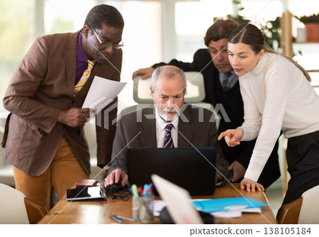 Team businessmen discusses business project while gathered around laptop in company office Team businessmen discusses business project while gathered around laptop in company office 138105184