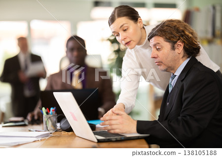 Woman leaned over to seated European man employee, helps with work and looks at laptop screen Woman leaned over to seated European man employee, helps with work and looks at laptop screen 138105188