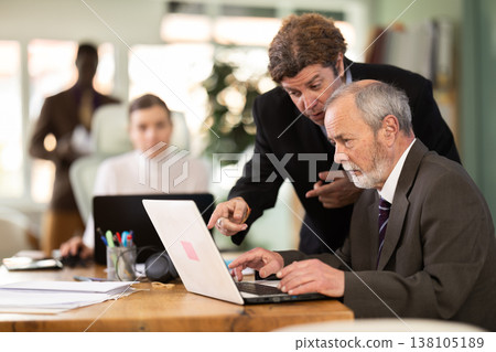 Man leaned over to seated mature male employee, helps with work and looks at laptop screen Man leaned over to seated mature male employee, helps with work and looks at laptop screen 138105189