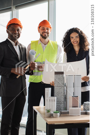 A diverse team of architects and engineers stand together in an office, reviewing a detailed architectural model of skyscrapers A diverse team of architects and engineers stand together in an office, reviewing a detailed architectural model of skyscrapers 138108315