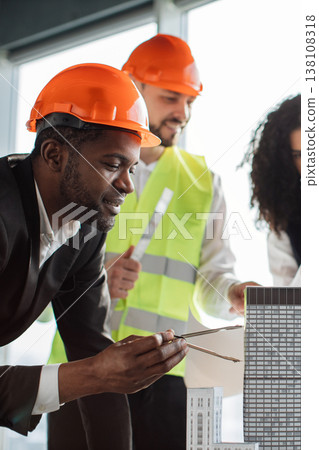 A diverse team of architects wearing hard hats and safety vests collaborates on a building project, examining blueprints and a miniature model of a skyscraper A diverse team of architects wearing hard hats and safety vests collaborates on a building project, examining blueprints and a miniature model of a skyscraper 138108318