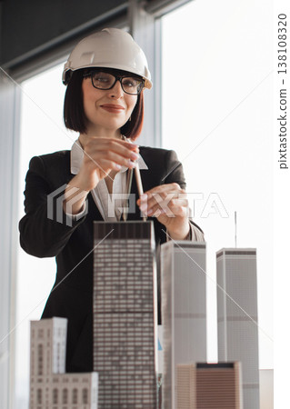 A woman wearing a hard hat and glasses meticulously adjusts a pencil over architectural models of skyscrapers in an office setting A woman wearing a hard hat and glasses meticulously adjusts a pencil over architectural models of skyscrapers in an office setting 138108320