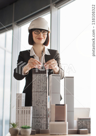 A smiling female architect wearing a hard hat and glasses uses a compass to measure architectural models of skyscrapers in an office setting A smiling female architect wearing a hard hat and glasses uses a compass to measure architectural models of skyscrapers in an office setting 138108322