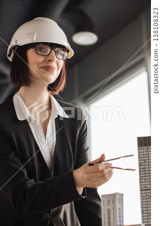 A female architect wearing a hard hat and glasses holds dividers near a building model, symbolizing construction and design planning A female architect wearing a hard hat and glasses holds dividers near a building model, symbolizing construction and design planning 138108323