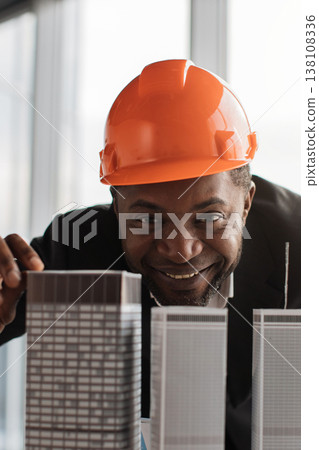 A smiling architect wearing an orange hard hat examines miniature skyscraper models in an office setting A smiling architect wearing an orange hard hat examines miniature skyscraper models in an office setting 138108336