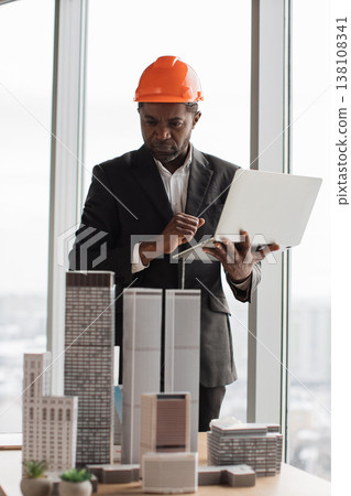 An African American architect wearing an orange hard hat examines a laptop while standing near architectural models of skyscrapers in an office An African American architect wearing an orange hard hat examines a laptop while standing near architectural models of skyscrapers in an office 138108341