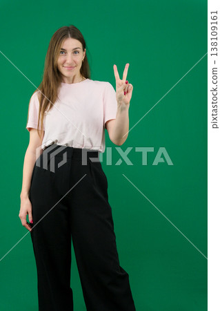 Pretty young woman showing peace gesture different emotions of a young girl on a green background chromakey beautiful facial features real people pink T-shirt white skinned European. black flared wide Pretty young woman showing peace gesture different emotions of a young girl on a green background chromakey beautiful facial features real people pink T-shirt white skinned European. black flared wide 138109161