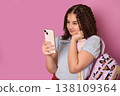 Young Girl with Curly Hair Engaged with Smartphone in Trendy Backpack Against Pink Background 138109364