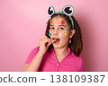 A Playful Girl with Colorful Candy Dots on Her Face, Enjoying Treats Against a Pink Background 138109387