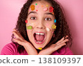 Joyful Expression of a Young Girl Surrounded by Colorful Candy on Her Face Against Pink Background 138109397