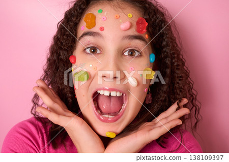 Joyful Expression of a Young Girl Surrounded by Colorful Candy on Her Face Against Pink Background 138109397