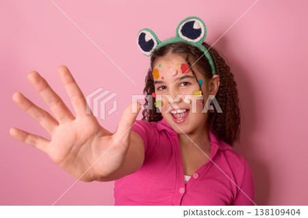 Joyful Girl Playfully Displaying Candy-Decorated Face Against a Vibrant Pink Background Joyful Girl Playfully Displaying Candy-Decorated Face Against a Vibrant Pink Background 138109404