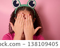 Child Hiding Face with Hands Wearing Fun Frog Headband Against a Soft Pink Background 138109405