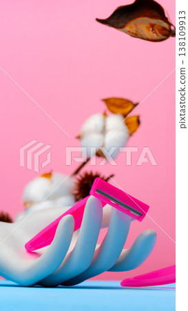 Hand holding pink razor with cotton flowers and leaves in the background on a colored surface Hand holding pink razor with cotton flowers and leaves in the background on a colored surface 138109913