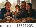 Family celebrating hanukkah: elderly female, young male, child, adult female lighting menorah with traditional foods 138110031
