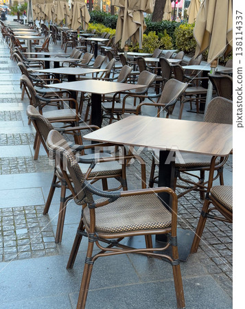 Empty outdoor cafe terrace with aligned wicker chairs and tables under folded umbrellas. Off peak dining setup, hospitality readiness, street seating arrangement, business downtime, urban service Empty outdoor cafe terrace with aligned wicker chairs and tables under folded umbrellas. Off peak dining setup, hospitality readiness, street seating arrangement, business downtime, urban service 138114337