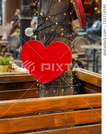 Red heart ornament attached to tree trunk with fairy lights above cafe terrace partition. Romance symbolism, seasonal decoration, date ambiance, hospitality styling, festive street dining atmosphere 138114348