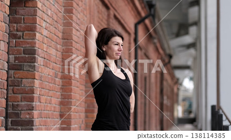 A woman performs a standing stretch, arms behind her back, leaning on a brick wall. This scene highlights the dedication to flexibility exercises and the importance of taking time for self-care. 138114385