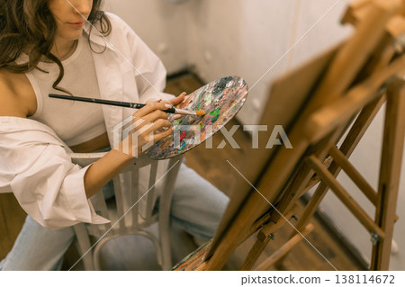 Young woman artist mixing paint on palette while painting on canvas at easel in art studio, creative hobby and painting process Young woman artist mixing paint on palette while painting on canvas at easel in art studio, creative hobby and painting process 138114672