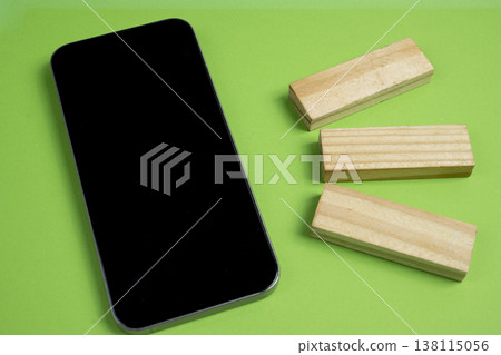 A sleek black smartphone lies next to three rectangular wooden blocks on a vibrant green surface, representing the intersection of modern technology and foundational building concepts A sleek black smartphone lies next to three rectangular wooden blocks on a vibrant green surface, representing the intersection of modern technology and foundational building concepts 138115056