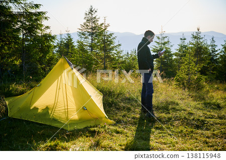 Camper stands near yellow tent, checking his phone in golden morning light. Campsite surrounded by lush evergreens and overlooks distant mountains. Camper stands near yellow tent, checking his phone in golden morning light. Campsite surrounded by lush evergreens and overlooks distant mountains. 138115948