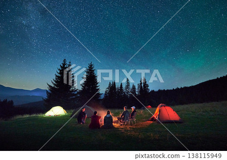 Group of friends hikers having a rest around campfire, surrounded by glowing tents under star-filled sky. Serene mountain backdrop and night sky create magical and peaceful atmosphere in wilderness. Group of friends hikers having a rest around campfire, surrounded by glowing tents under star-filled sky. Serene mountain backdrop and night sky create magical and peaceful atmosphere in wilderness. 138115949
