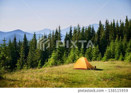 Bright orange tourist tent on grassy hillside, surrounded by lush green pine trees. Breathtaking view of distant blue mountains under clear sky, creating idyllic and serene camping spot. Bright orange tourist tent on grassy hillside, surrounded by lush green pine trees. Breathtaking view of distant blue mountains under clear sky, creating idyllic and serene camping spot. 138115959