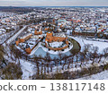 Aerial view of a medieval brick castle with a water filled moat in a snowy European town. Picturesque winter landscape featuring historical architecture, a church spire, and frosted rooftops. 138117148