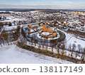 Aerial view of a medieval brick castle with a water filled moat in a snowy European town. Picturesque winter landscape featuring historical architecture, a church spire, and frosted rooftops. 138117149
