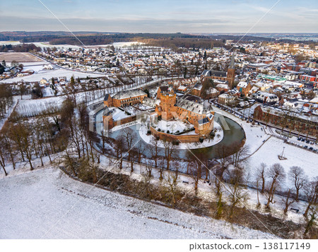 Aerial view of a medieval brick castle with a water filled moat in a snowy European town. Picturesque winter landscape featuring historical architecture, a church spire, and frosted rooftops. Aerial view of a medieval brick castle with a water filled moat in a snowy European town. Picturesque winter landscape featuring historical architecture, a church spire, and frosted rooftops. 138117149