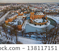 Aerial view of a medieval brick castle with a water filled moat in a snowy European town. Picturesque winter landscape featuring historical architecture, a church spire, and frosted rooftops. 138117151