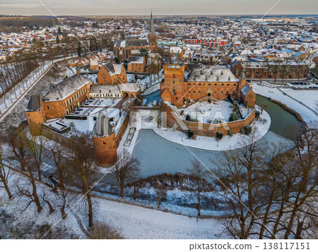 Aerial view of a medieval brick castle with a water filled moat in a snowy European town. Picturesque winter landscape featuring historical architecture, a church spire, and frosted rooftops. Aerial view of a medieval brick castle with a water filled moat in a snowy European town. Picturesque winter landscape featuring historical architecture, a church spire, and frosted rooftops. 138117151