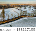 Aerial view of a snowy Dutch landscape at sunset. A small canal winds through frost-covered fields and rows of bare trees, with a charming European village and church spire on the golden horizon. 138117155