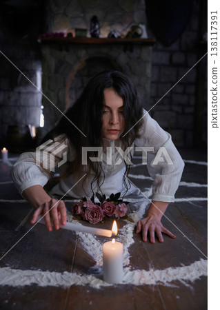 Woman with dark hair meticulously outlines sacred symbol on floor surrounded by candles and flowers Woman with dark hair meticulously outlines sacred symbol on floor surrounded by candles and flowers 138117391