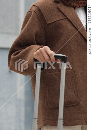 Close-up of a woman in a brown wool coat holding the handle of a suitcase. The shot expresses calm readiness, autumn travel mood, and refined minimalism in neutral tones. Close-up of a woman in a brown wool coat holding the handle of a suitcase. The shot expresses calm readiness, autumn travel mood, and refined minimalism in neutral tones. 138119014