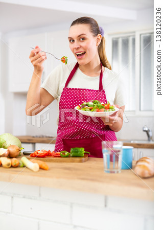 Smiling young woman eating vegetable salad in home kitchen 138120236